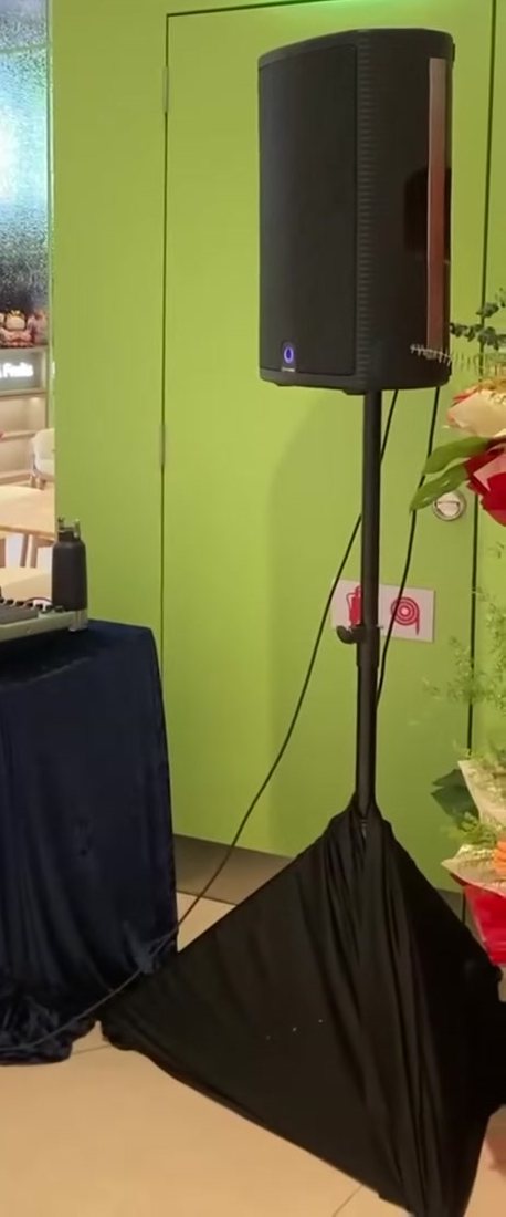 Powered PA speaker on a tripod stand with a black skirt at an indoor retail opening, mixer on a draped table behind