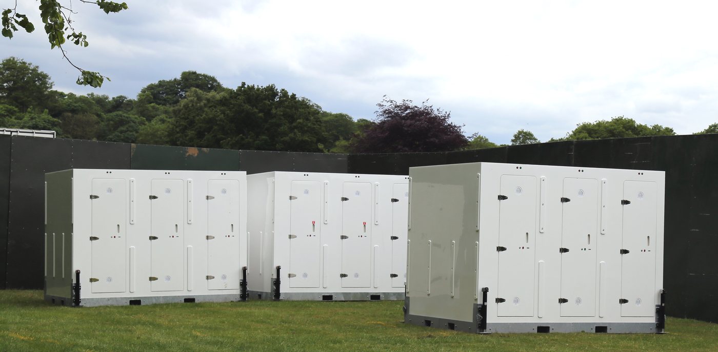 Three banks of white portable toilet cabins standing on grass at an outdoor event field behind temporary fencing